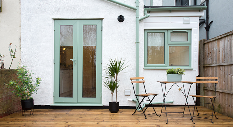 A general exterior view of a back garden patio area with wood decking, potted plants, Dragon palm tree, metal table and two chairs pale pastel sage green patio doors, window and drainpipe